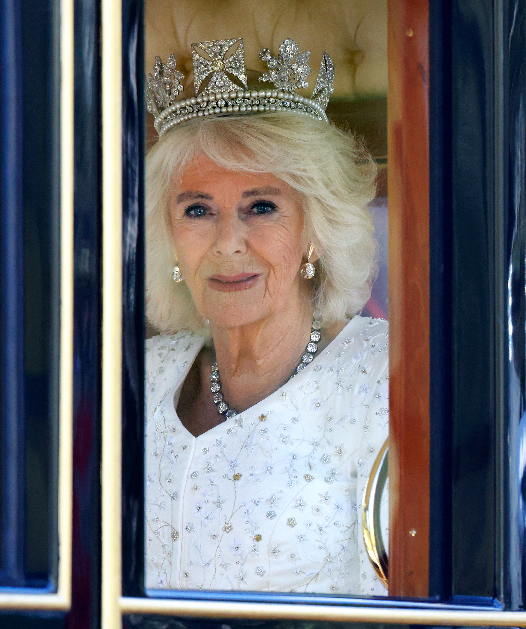 Queen Camilla wearing a diamond tiara and white dress riding in a carriage to the state opening of parliament in 2023