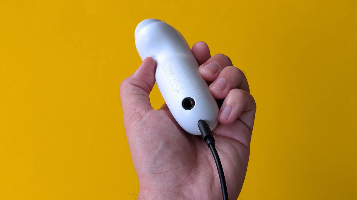 A hand holds a small white XBOX Adaptive Joystick against a bright yellow background. A black cable is plugged into the bottom, and faint manufacturer markings are visible on the casing.