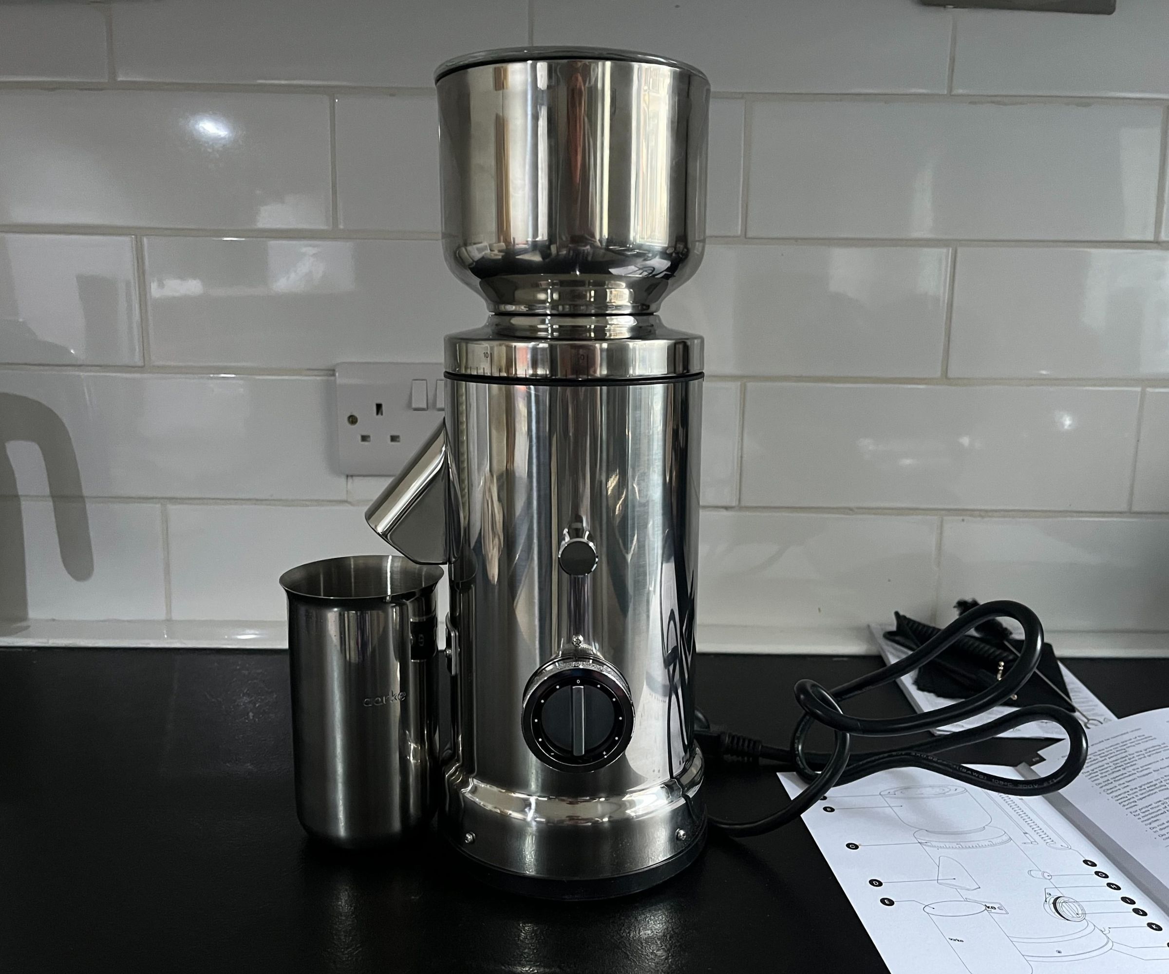 A tall stainless steel coffee grinder on a black counter top in front of a white subway tile wall