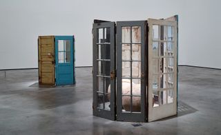 Two sculptures in the space. Closest (right) is a front-side view of three grey doors adjoined to each other with eight windows in each. Back (left) are two doors joined together, one brown with no windows and one green with four windows.