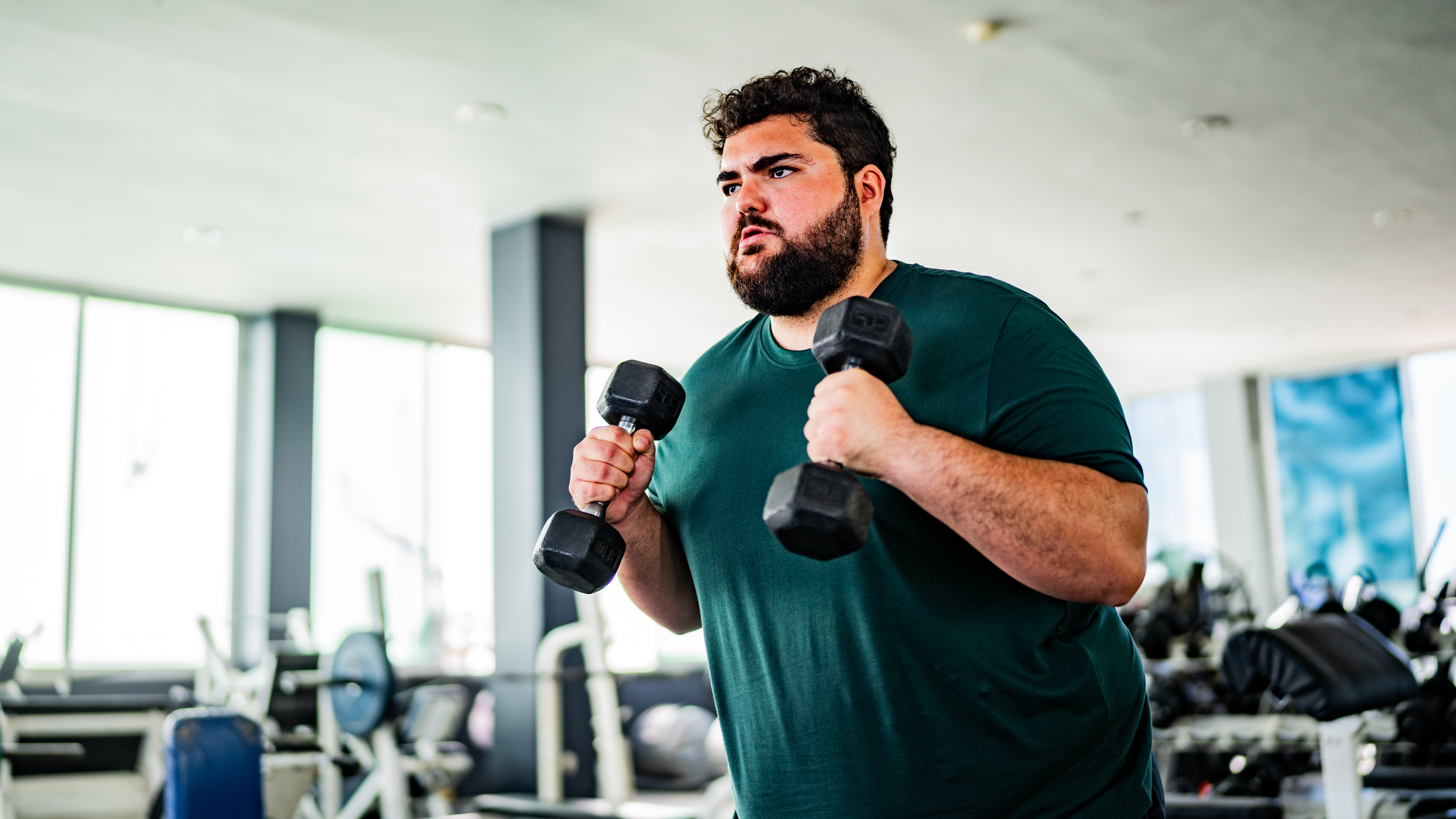 A man stands in a gym looking determined, holding two dumbbells at chest height.