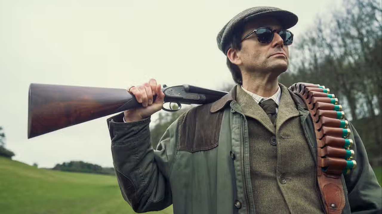 Lord Tony Baddingham standing in a field with a rifle cocked on his shoulder in Disney's Rivals TV show