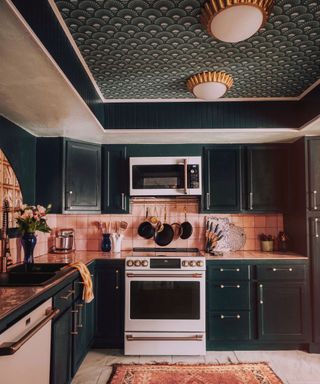 Kitchen with dark green and gold art deco inspired wallpaper, matching dark green cabinets, pink wall tiles, marble flooring, pink patterned rug