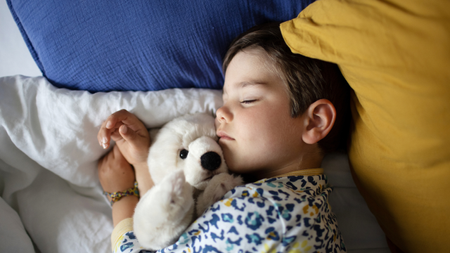 A young child lies asleep on his side in bed while cuddling a white fluffy teddy bear.