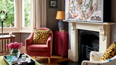 a striped living room with traditional fireplace, large artwork box hiding a TV, a small pink armchair with a leopard print pillow and a series of eclectic side tables in the bay window and alcove