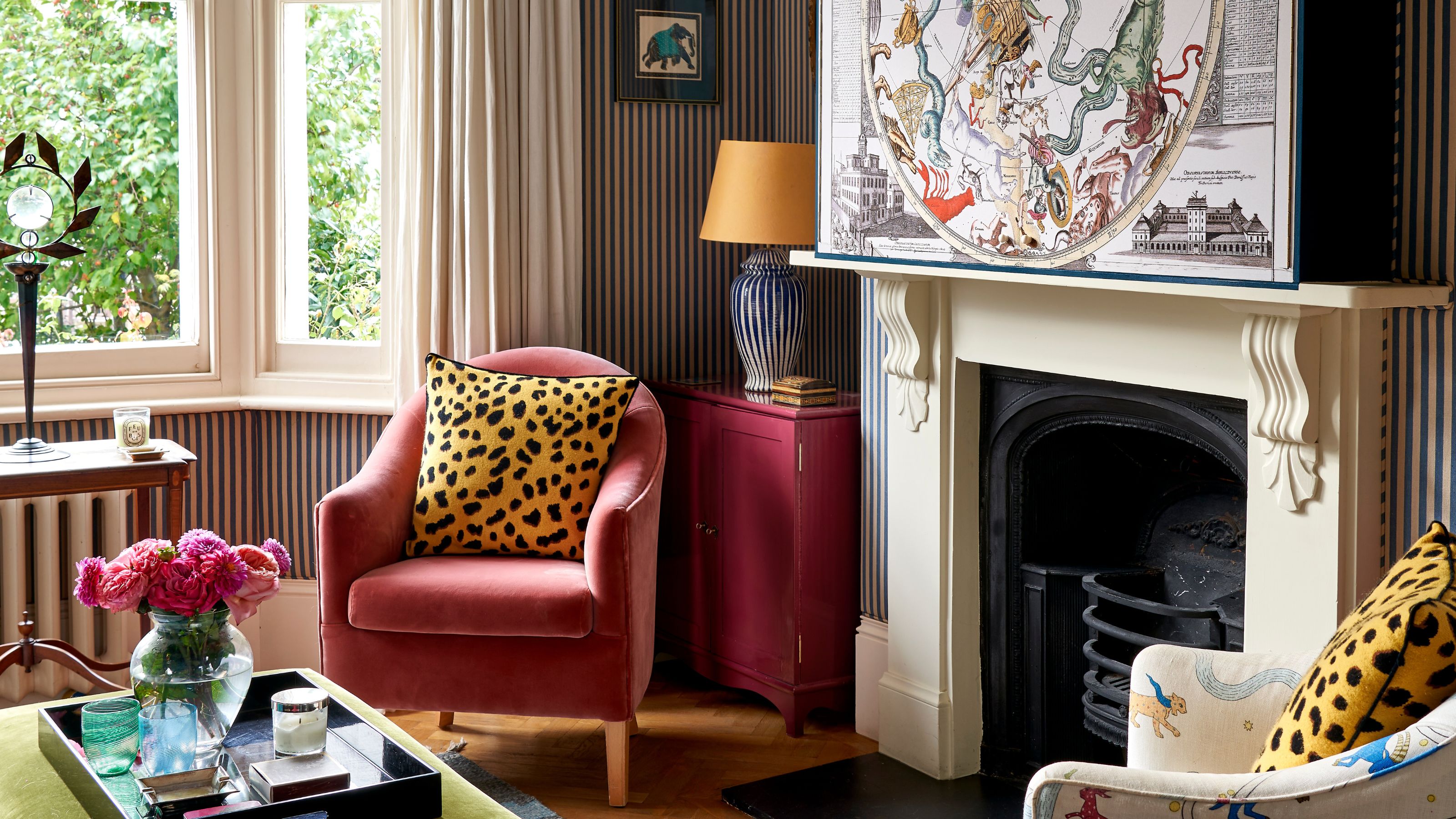a striped living room with traditional fireplace, large artwork box hiding a TV, a small pink armchair with a leopard print pillow and a series of eclectic side tables in the bay window and alcove