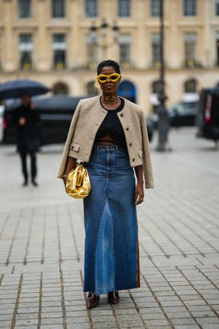 woman in a maxi denim skirt, black top, tan jacket, and yellow sunglasses