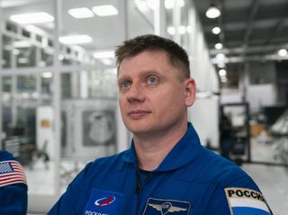 a man wearing a blue jumpsuit stares up into the distance. He sits in a white room with many glass panels.