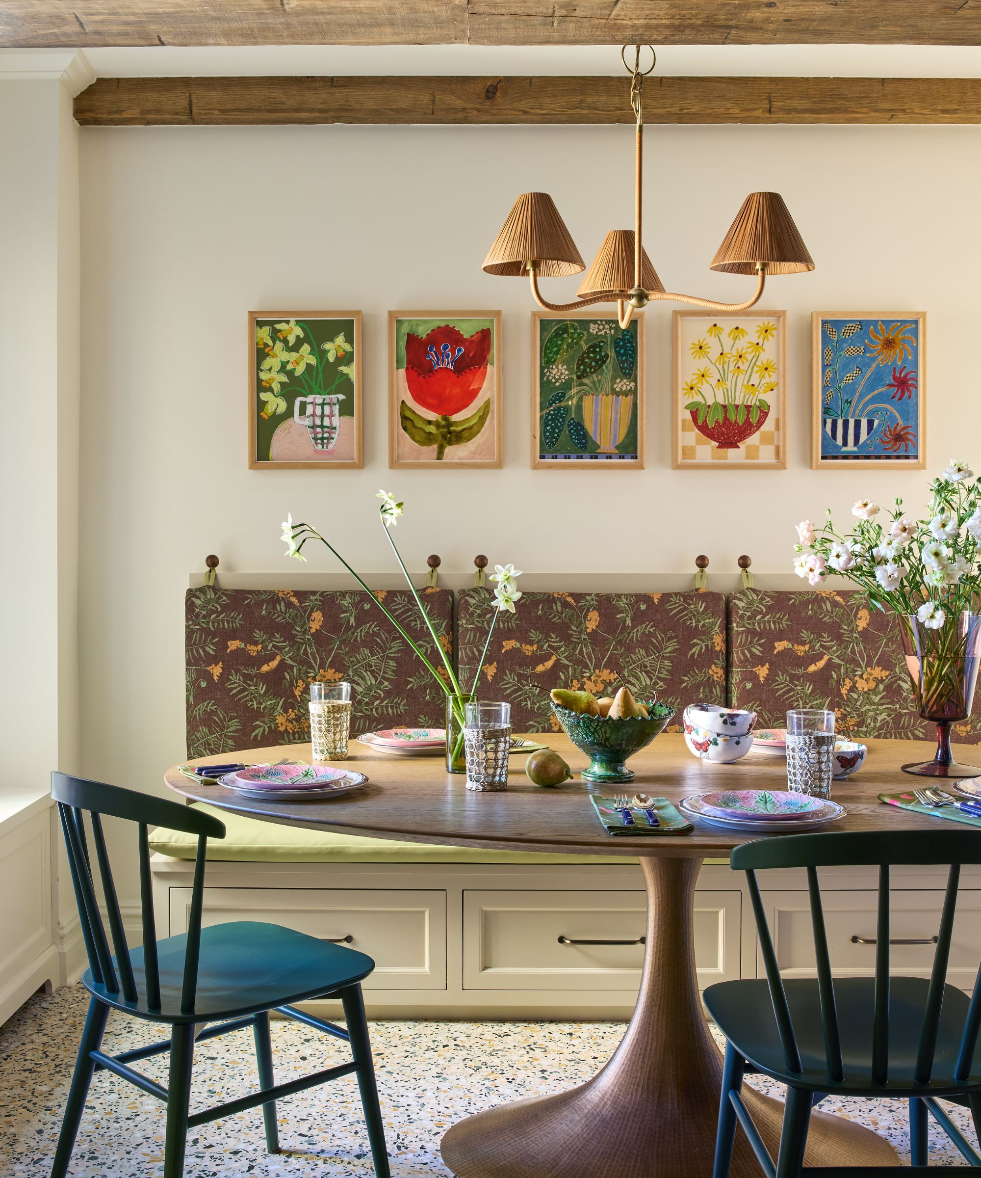 A breakfast nook with neutral walls and a collection of colorful artworks. A round dining table with two chairs and banquette seating with brown floral upholstery.