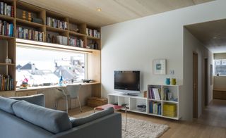 Interior view of a living area at the Oslo family house featuring white walls, wood flooring, a wood panelled ceiling with spotlights, a blue sofa, an off-white coloured rug, a small table, wooden and white shelving units with books and other items, a tv and a window with a light coloured chair in front
