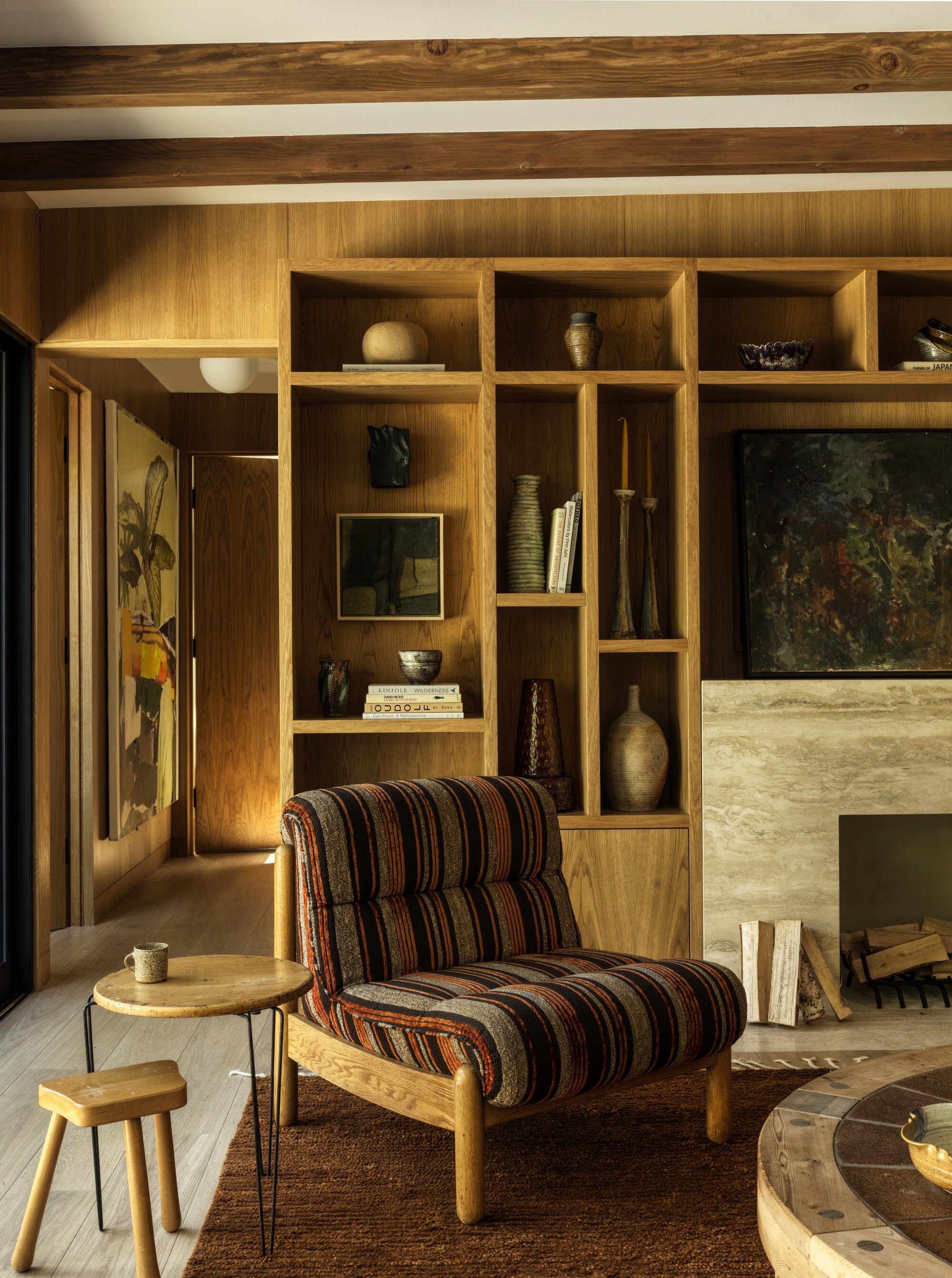 A cozy dining room with built-in wooden shelving and a marble fireplace. There are wooden beams on the ceiling and the furniture in the room is also wooden. The accent chair has a striped pattern and a tobacco colored rug underneath.