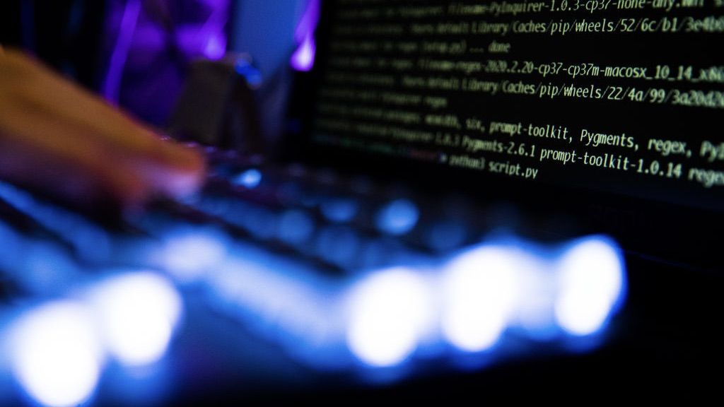 A person types at a backlit keyboard in front of a code terminal