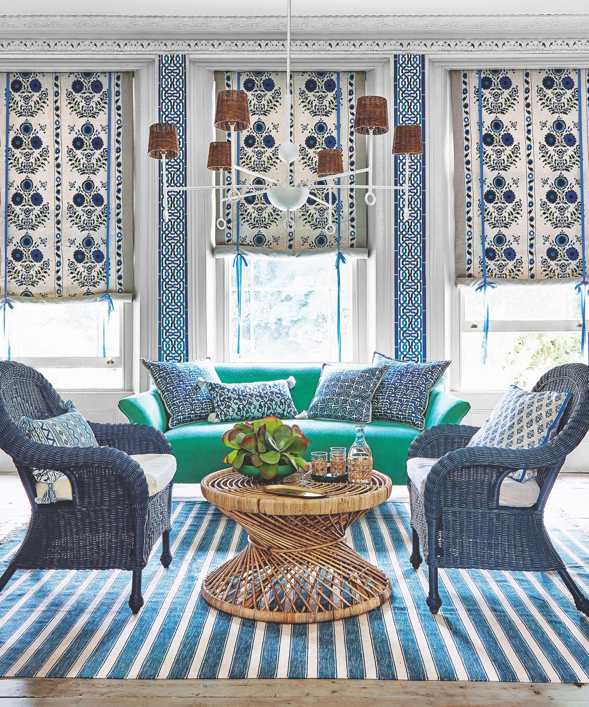 A seating area in a maximalist living room, with a striped blue and white rug, a circular wicker table in the center, with two matching wicker chairs with white and blue patterned cushions to either side, and a bright green couch with matching cushions behind. Behind the couch are three large windows with blue and white patterned blinds, which are seen half closed. There is a large white statement lamp hanging in the center of the room with small wicker shades. The floor, seen in the foreground, is light wood.