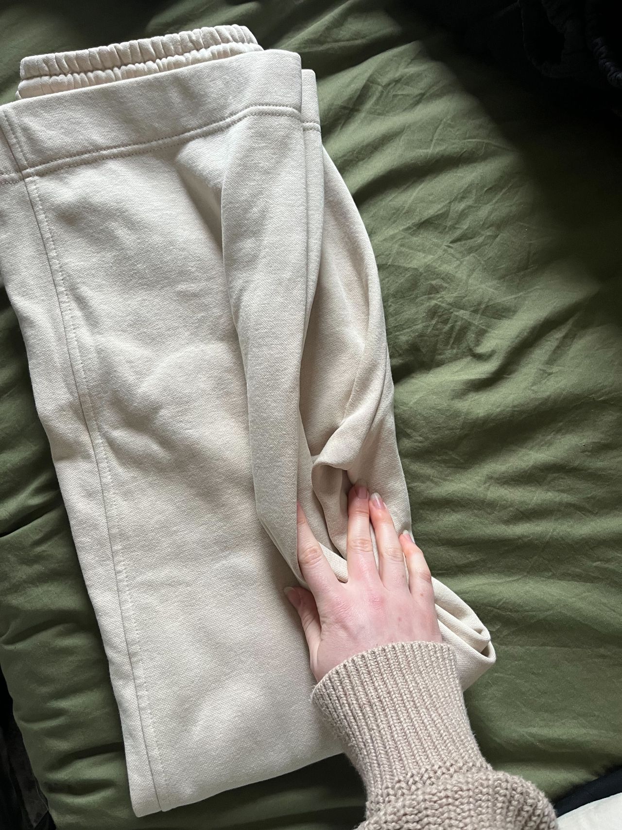 A pair of cream sweatpants folded in half on a green bed, a hand showing the folding technique