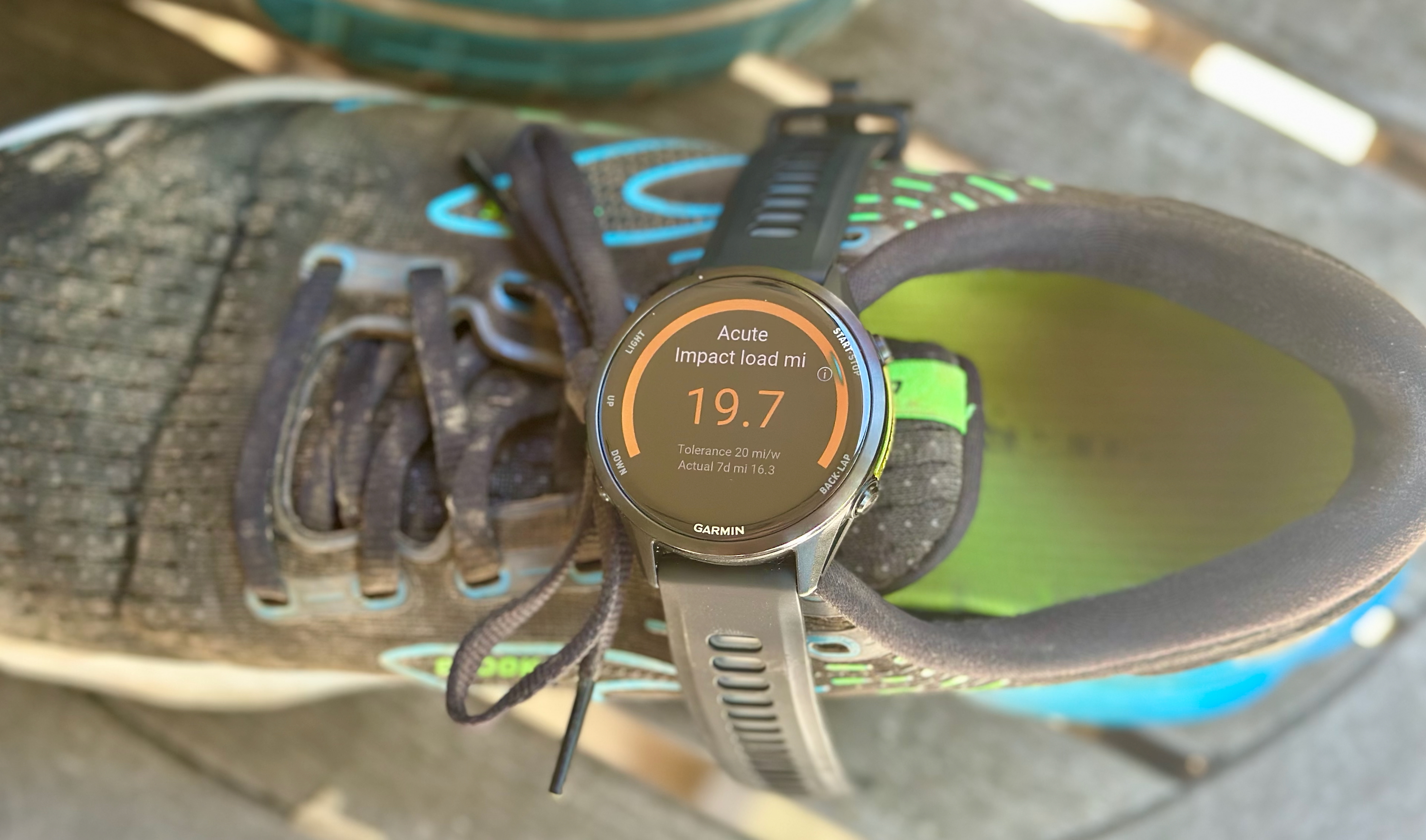 The Garmin Forerunner 970 sitting atop a running shoe, showing the Acute Impact Load Miles widget with the runner's current mileage compared to their weekly tolerance and actual weekly mileage.