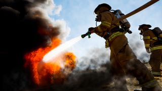Firefighters in a fire protection suit wearing firefighter helmet with breathing device and holding fire hose is extinguishing a burning house fire that is putting off excessive heat and smoke.