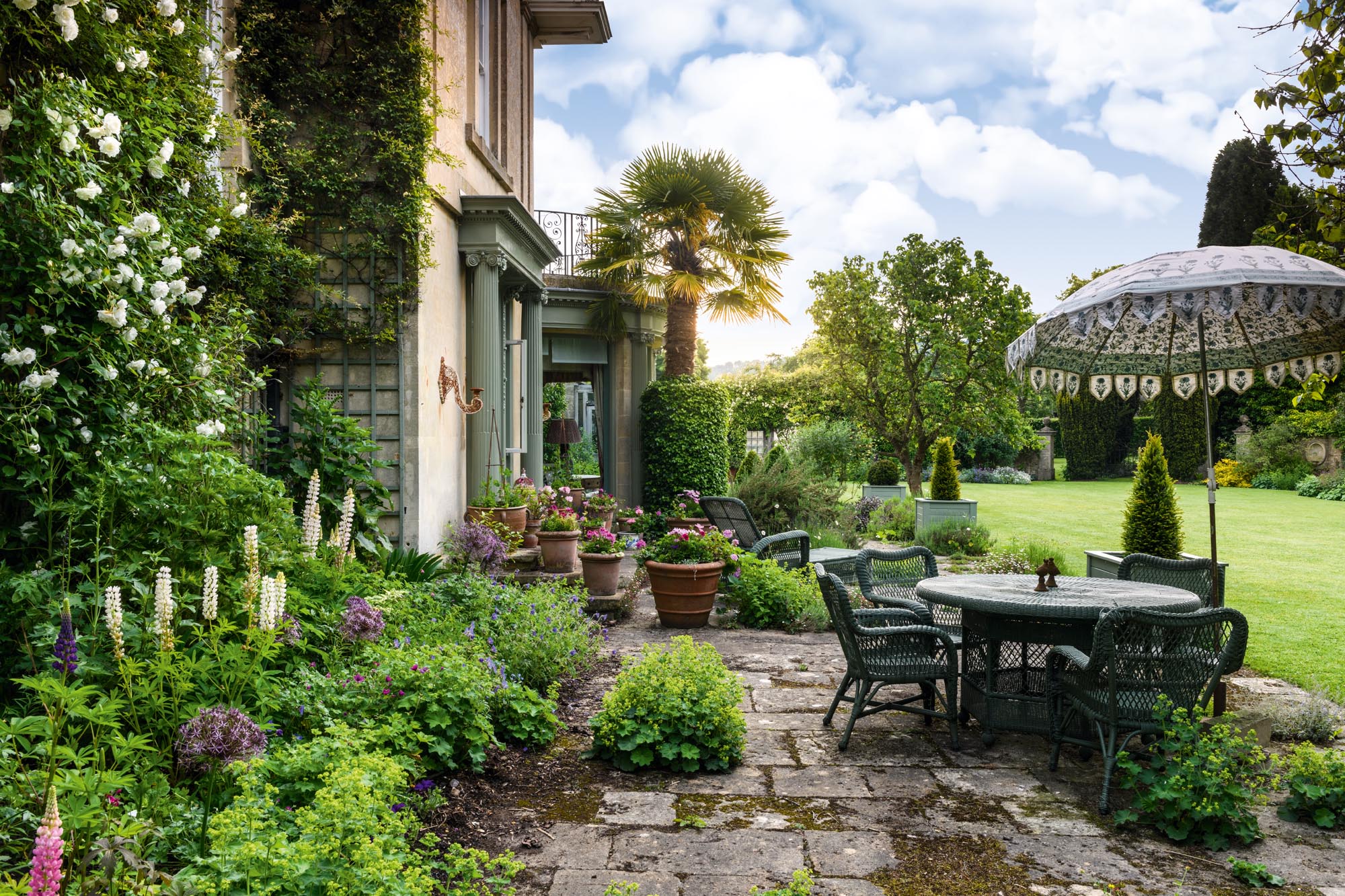 The terrace, with the vigorous double white Rosa ‘Climbing Iceberg’, lupins and geraniums and Alchemilla mollis between the flagstones, sets the tone for the garden. Raymill, Wiltshire.