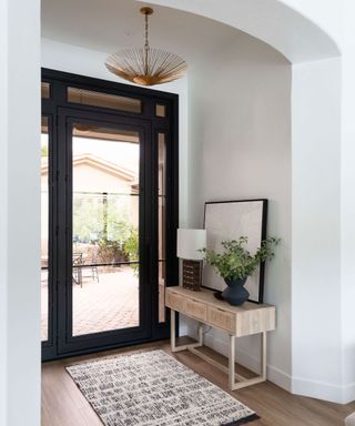 Lofty entryway with textured rug, slimline wooden console with framed, leaning mirror placed behind table lamp and indoor plant