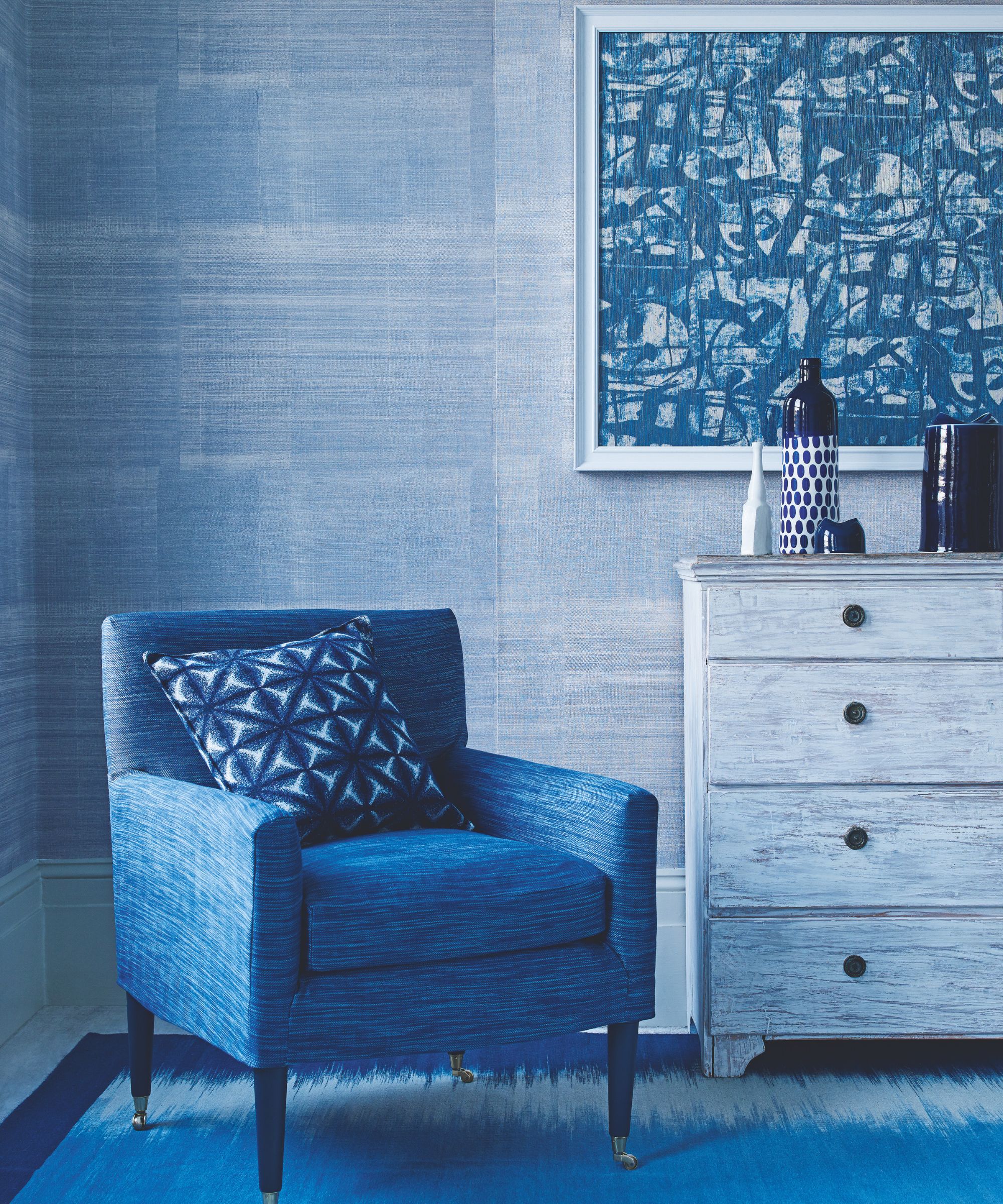 A blue bedroom with a blue rug, blue upholstered chair with blue patterned cushion, blue dresser with bottle and ornaments on top and a blue abstract artwork above in a white frame. The wall is also blue wallpaper.