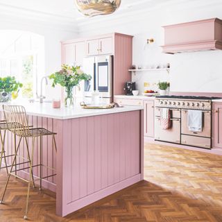 pink and white kitchen with herringbone floor, brass bar stools, chrome range cooker, open plan shelving, plants, vases of flowers