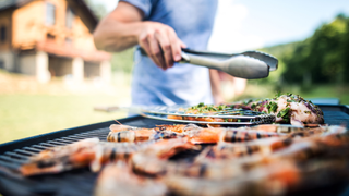 Image depicts meat being cooked on BBQ and man with tongs.