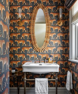 Bathroom space with bold tiger wallpaper with black background, traditional sink with exposed piping, oval mirror with bamboo style edging
