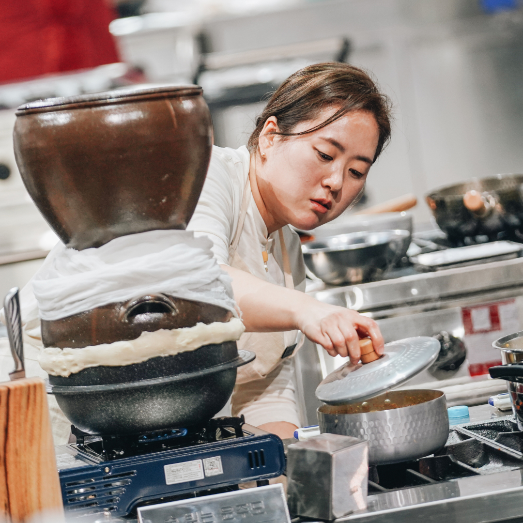 A female chef nicknamed Brewmaster Yun lifts a lid on a small pot while standing at a workstation with a traditional liquor distilling pot, in 'Culinary Class Wars' season 2.