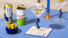 A blue tablecloth with a pair of martini glasses with olive picks, a taper candle, a can of olives, a can of sparkling water, and a bowl of olives