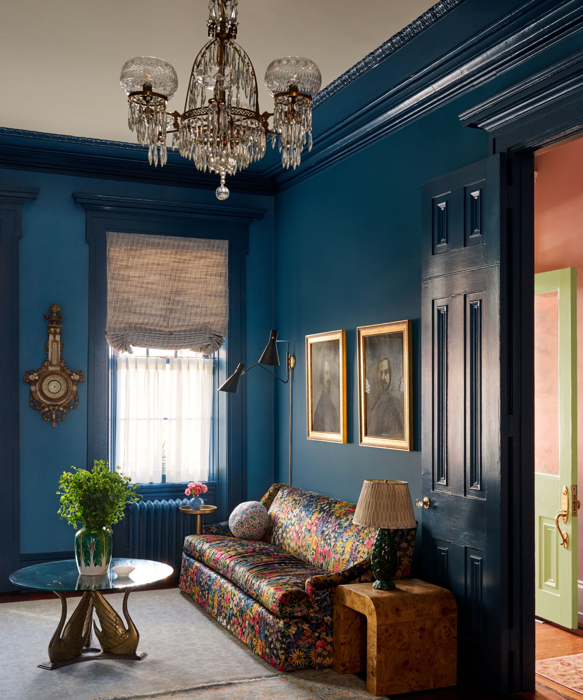Living room with teal walls, a multi-colored floral tapestry sofa, framed portraits, and a glass swan coffee table.
