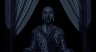 Lily Rose Depp's character (Ellen) from Nosferatu stands under a window with curtains peeled back on either side. In the middle of her face and the top half of her body is the shadow of a gnarled hand.