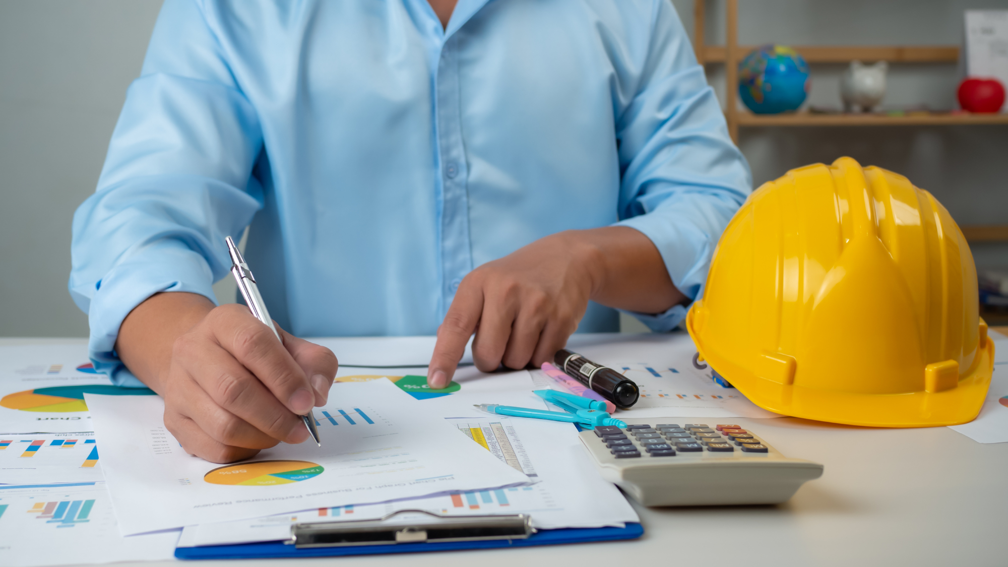 male in blue shirt working at desk making marks on graphs, with calculator and hard hat on table