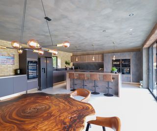 large open plan kitchen diner with dark grey suede effect kitchen units, combined with wood finishes, island with brown leather bar stools and large natural wood organically shaped table