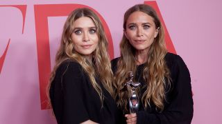 Mary-Kate and Ashley Olsen both wear black and stare away from the camera. The background is pink and Mary-Kate is holding a statue. 