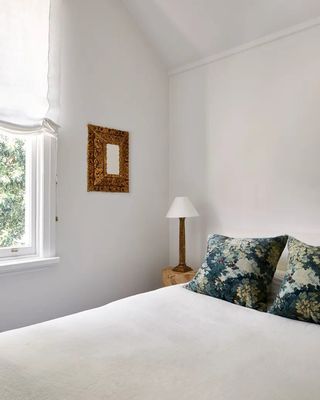 bright light bedroom with white walls, relaxed roman blinds over window, an antique gold mirror onthe wall, a burl bedside with a table lamp, and verdure print pillows on the bed