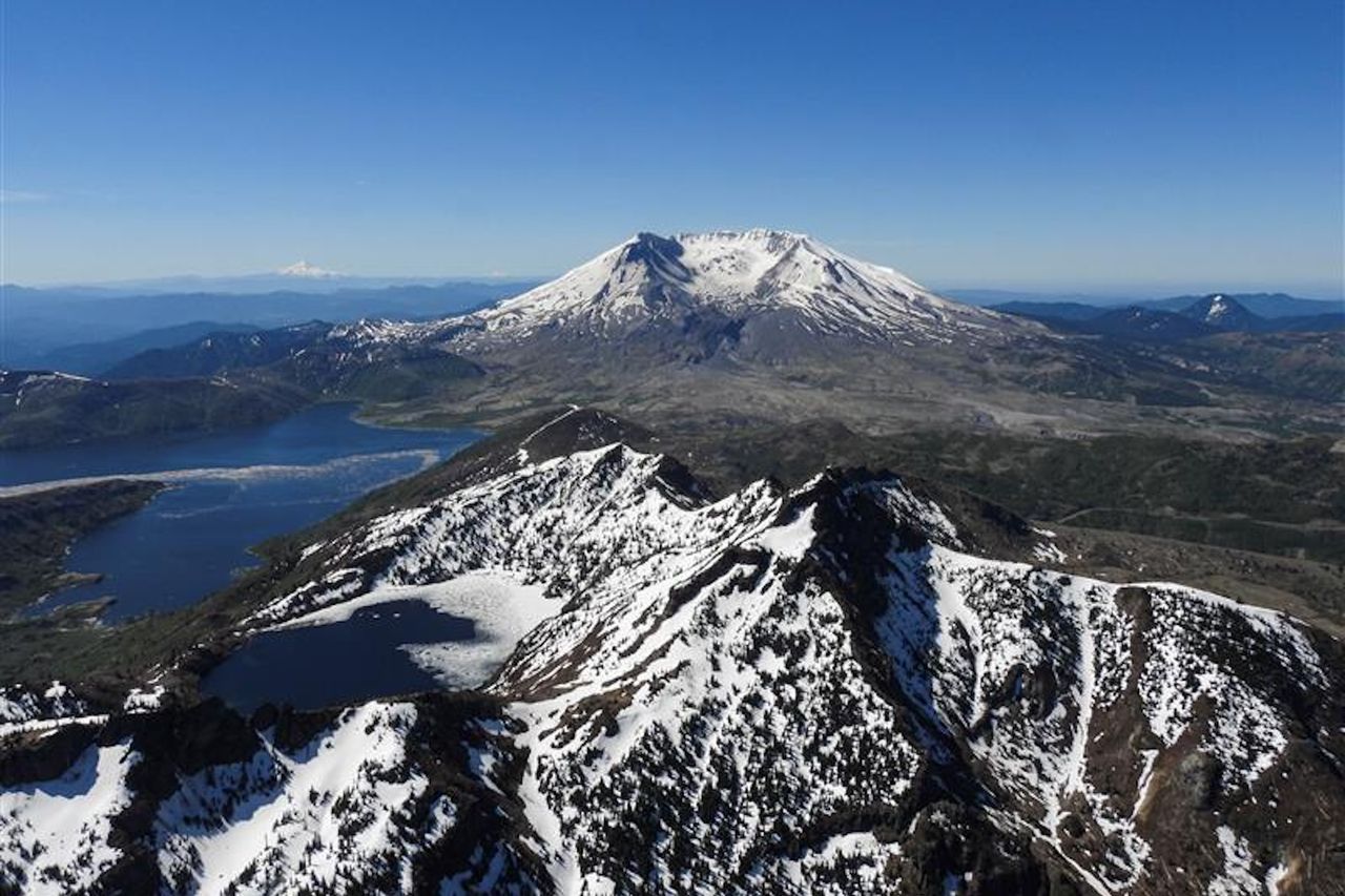 A snow-covered mountain is flanked by a large blue lake to the left and additional snowy mountainous terrain in the foreground.