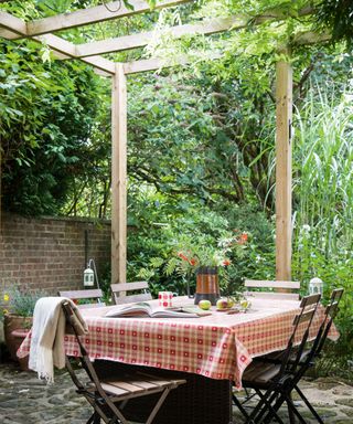 Outdoor setting with wooden pergola, covered with greenery, square cafe table with four chairs, red gingham tablecloth