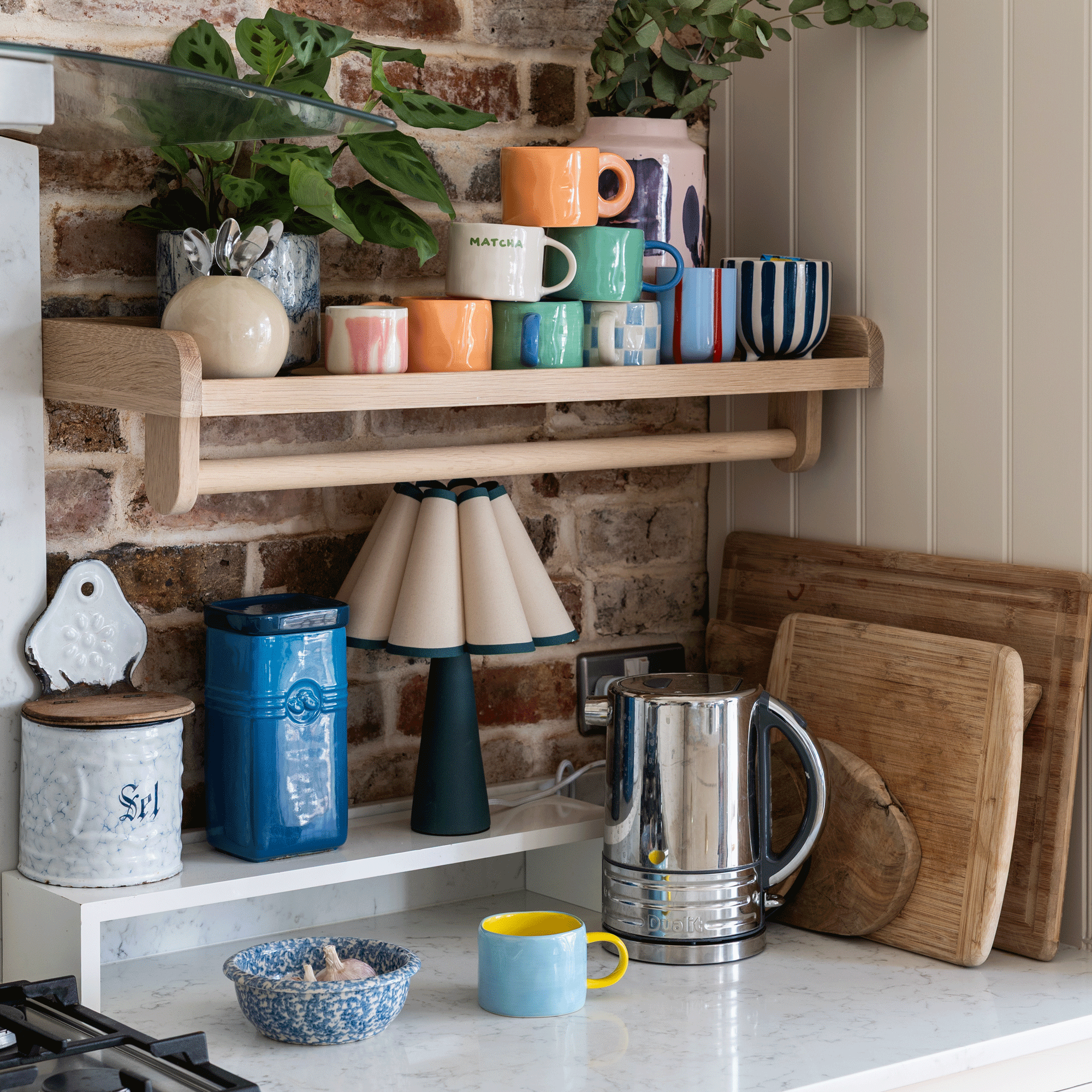 a tea station in a kitchen with a shelf for colourful mugs and kettle a lamp and a tea caddy