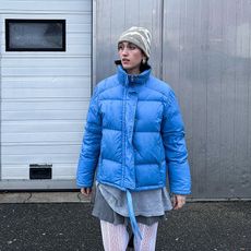 leonie wears gray outfit and white tights with a blue puffer jacket over everything.