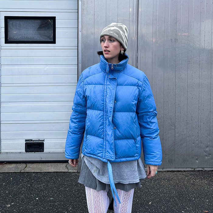 leonie wears gray outfit and white tights with a blue puffer jacket over everything. 