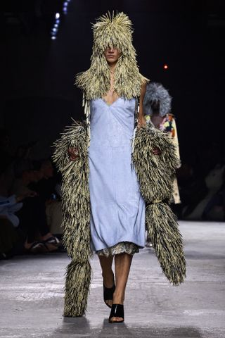 A model at Bottega Veneta on the runway during the spring/summer 2025 season wearing a yellow-and-blue string hat and shawl with a light blue slip dress and mules.