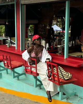 Woman wearing a red baseball cap, a striped bikini top, a white button down, and white linen pants.