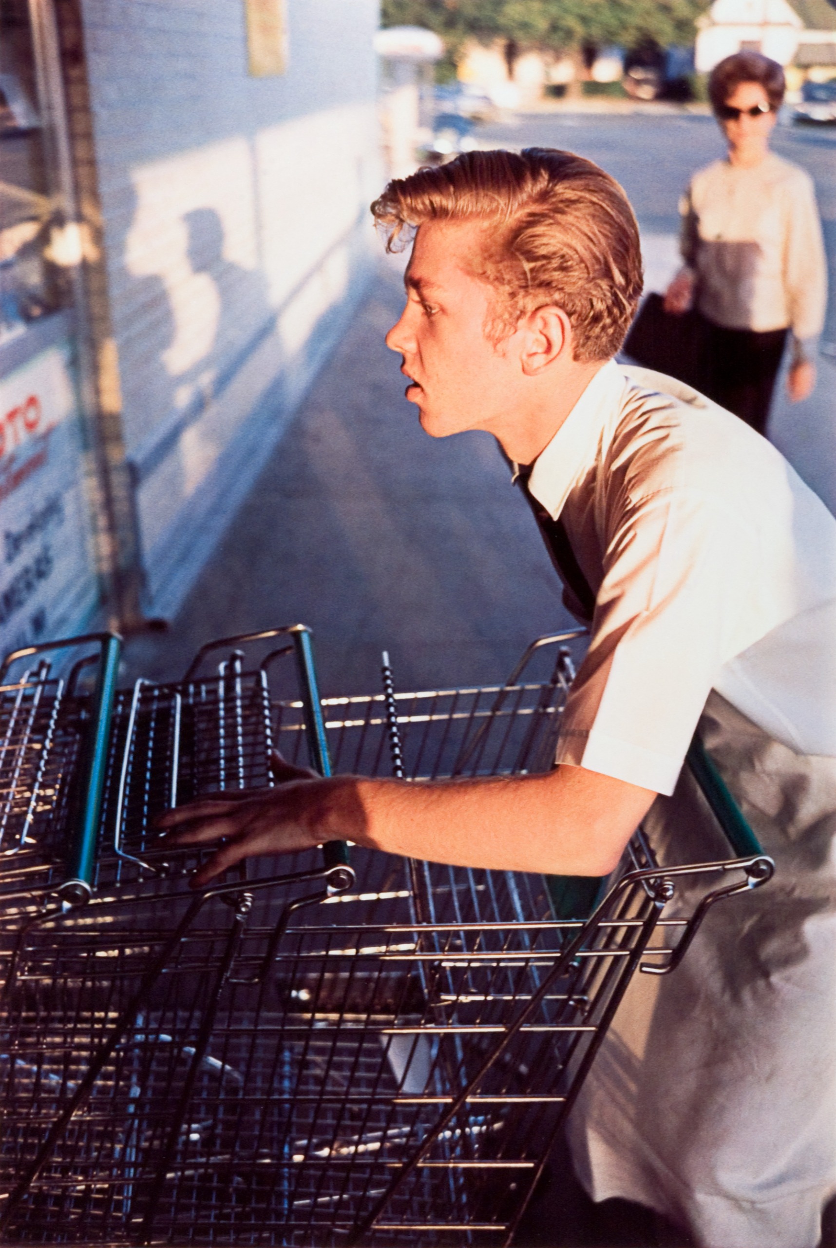 A color photograph of a young man leaning over a row of stacked shopping trolleys outside a store, with a woman walking behind him.