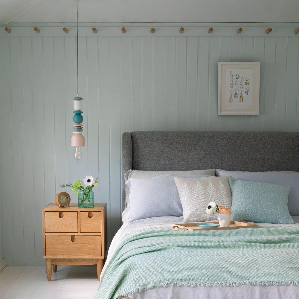 Bedroom with panelled wall, peg rail, grey fabric headboard and wooden side table.