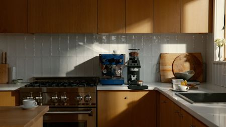 Image of a blue La Marzocco coffee machine next to a black grinder from the brand on a white countertop in a kitchen with wooden cabinetry, white backsplash, and silver appliances.s