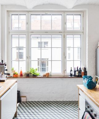 A white kitchen with a large grid window, a windowsill with herbs and bottles on, two wooden kitchen counters to the left and right, and green, black, and white geometric flooring