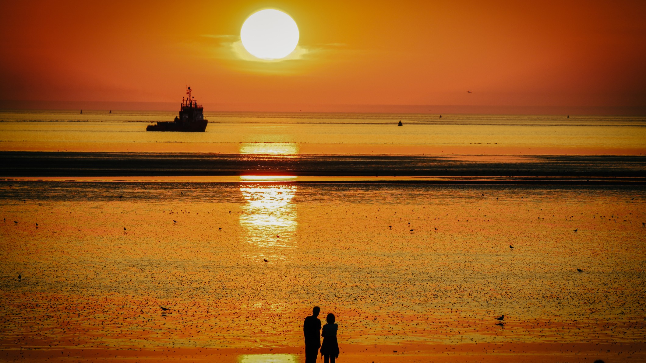 View of a sunset over the ocean with a boat and a couple standing on the beach.
