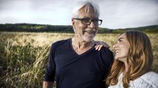 A father and his adult daughter smile at each other while standing in a field.