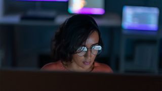 Female Java developer working at a computer station in low light with screen reflecting on glasses. 