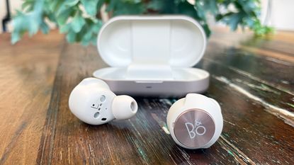 Bang & Olufsen Beoplay EQ review, image of the earbuds on a wooden table, one has the B&O logo facing the camera, with the case in the background