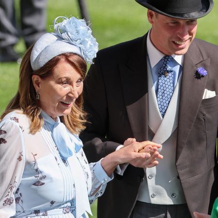 Carole Middleton wearing a pale blue floral dress and matching headband with a flower holding hands with Prince William, who is wearing a suit and top hat while standing in grass at Royal Ascot 